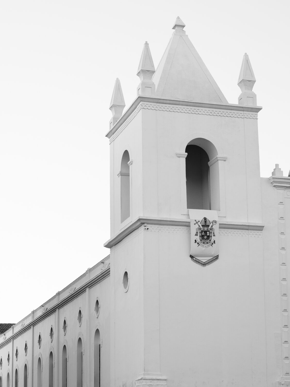 Torre branca da igreja com pináculos e o estandarte da diocese, sob céu claro.