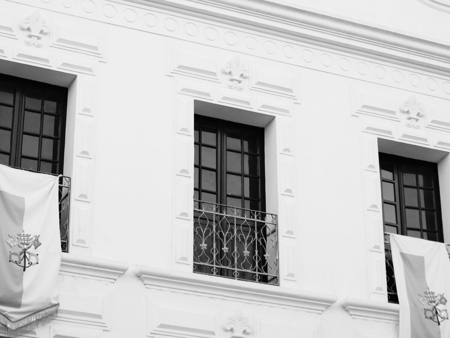 Janelas de madeira escura com grades de ferro e bandeiras da Santa Sé na fachada branca.