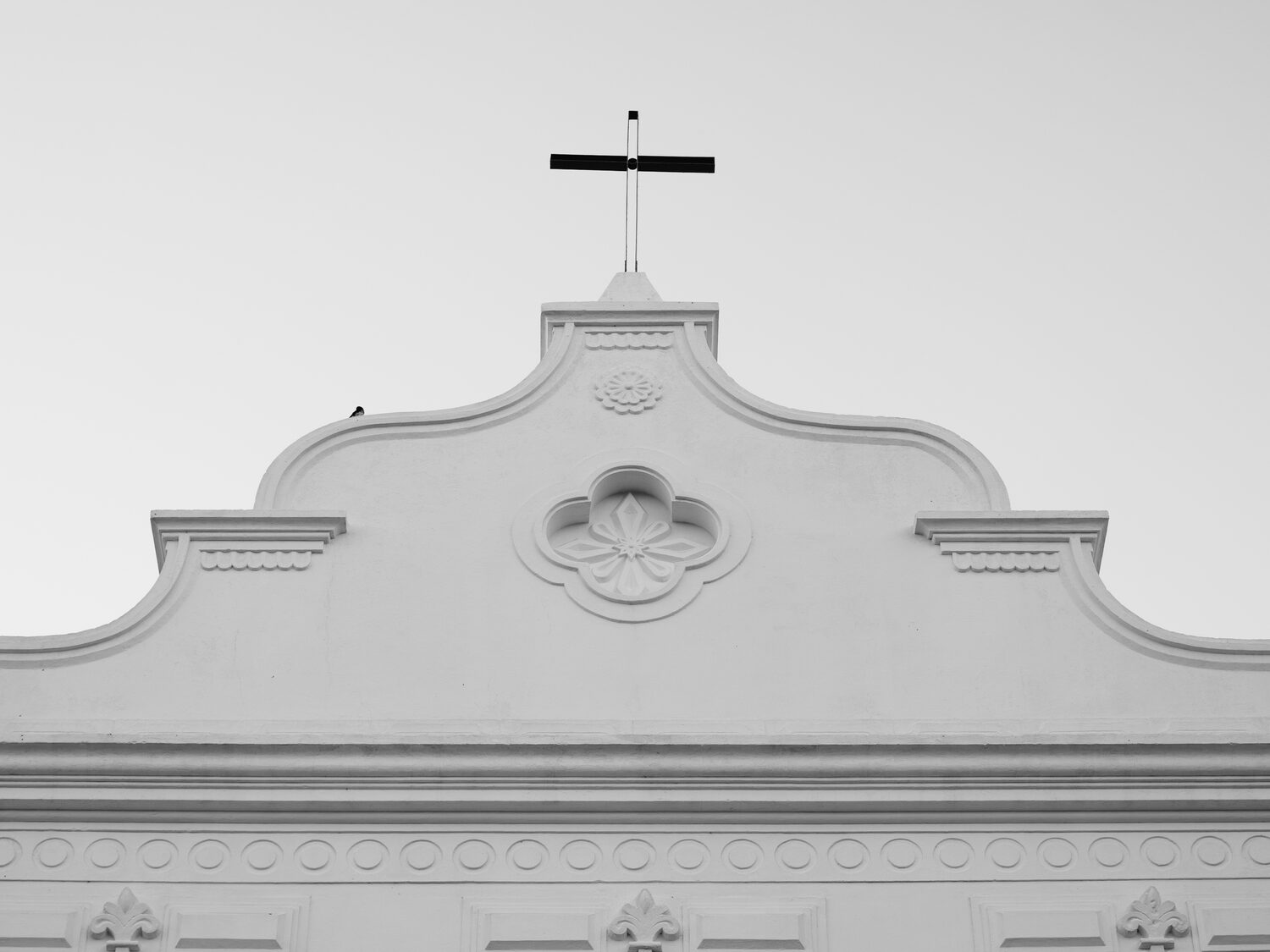 Cruz escura no topo do frontão branco contra o céu claro.