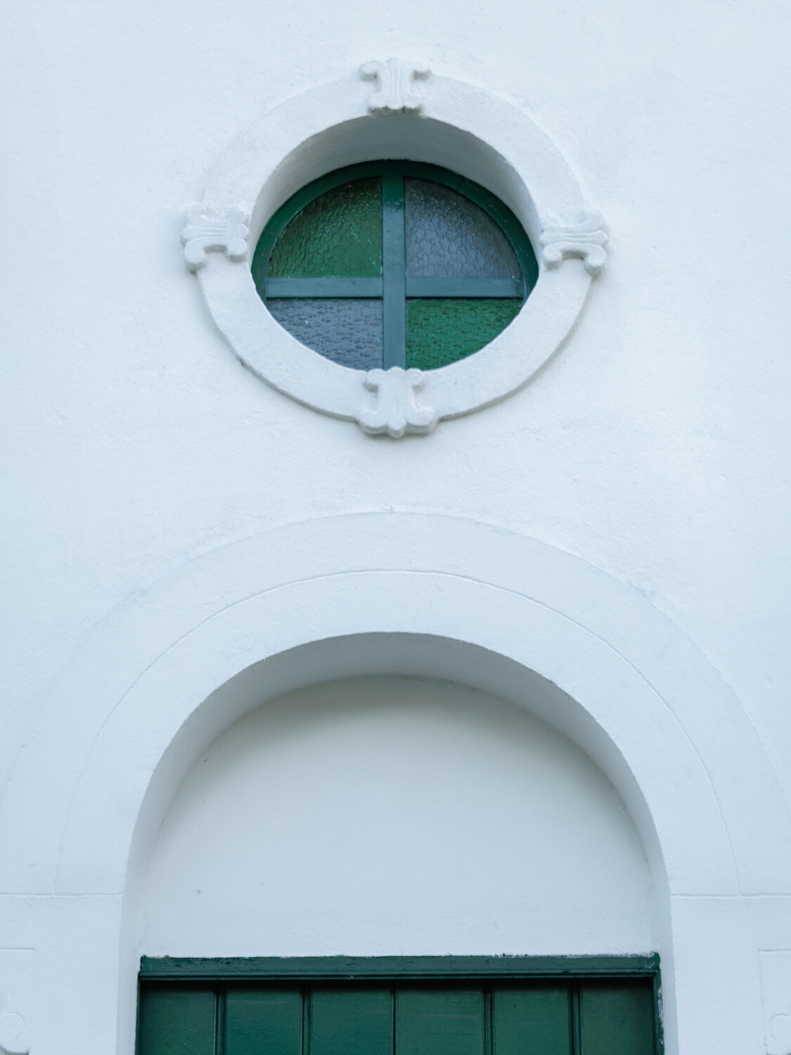 Óculo circular com vitral verde em forma de cruz, sobre uma porta em arco na parede branca.